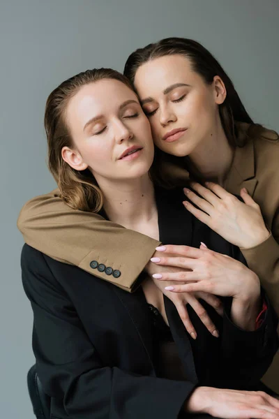 Чувственная пара лесбиянок в бежевых и черных пиджаках обнимающихся с закрытыми глазами, изолированными на сером — стоковое фото