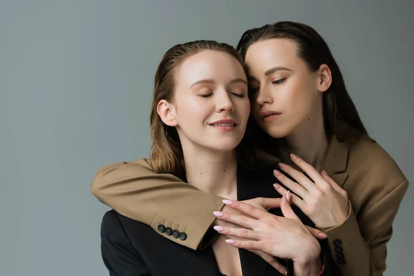 Mulher morena em blazer bege abraçando namorada lésbica sorrindo com olhos fechados isolados em cinza — Fotografia de Stock