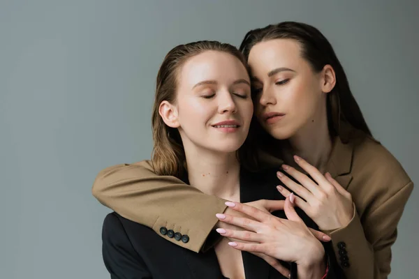 Morena mujer en chaqueta beige abrazando novia lesbiana sonriendo con los ojos cerrados aislados en gris — Stock Photo