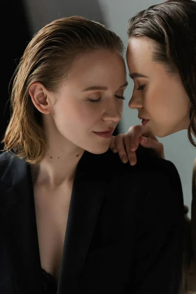 Morena lesbiana mujer cerca de joven novia con los ojos cerrados sobre fondo gris - foto de stock