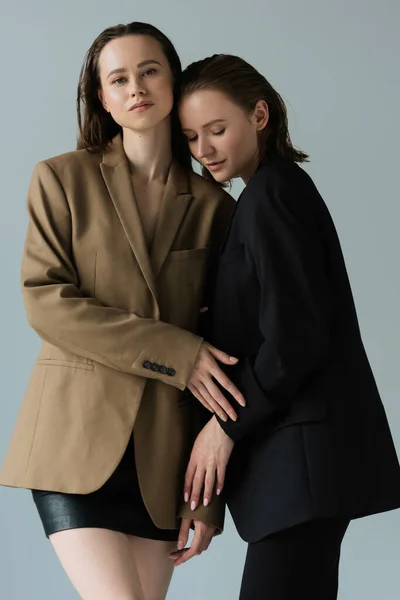 Brunette femme en blazer beige et jupe noire regardant la caméra près partenaire lesbienne isolé sur gris — Photo de stock