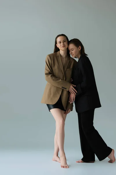 Full length of barefoot woman in blazer and skirt looking at camera near lesbian girlfriend in suit on grey background — Stock Photo