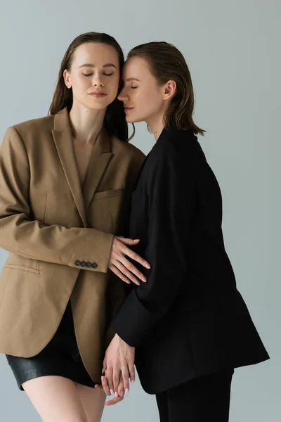 Mujeres lesbianas jóvenes en blazers negros y beige de pie con los ojos cerrados aislados en gris - foto de stock