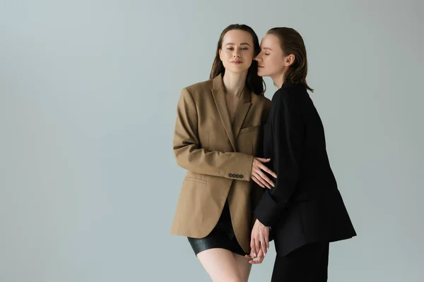 Bonita mujer lesbiana en traje negro cogida de la mano con novia en blazer beige aislado en gris - foto de stock