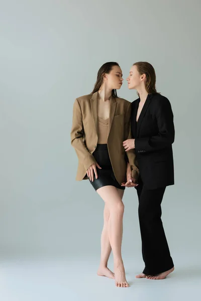 Mujer en traje negro y su pareja lesbiana en blazer y falda tomados de la mano y mirándose el uno al otro sobre fondo gris - foto de stock