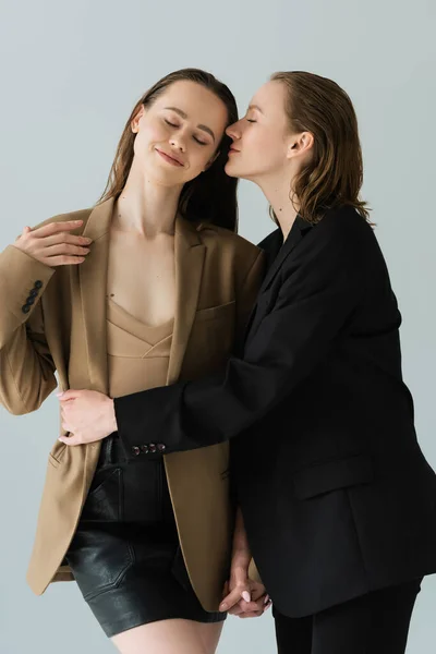 Young woman in black blazer embracing girlfriend standing with closed eyes isolated on grey — Stock Photo