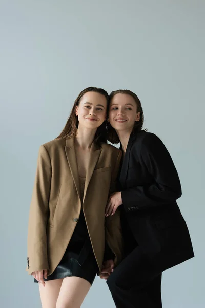 Young and happy lesbian partners in blazers holding hands isolated on grey — Stock Photo