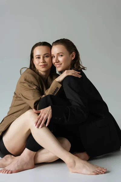 Fröhliche lesbische Partnerinnen in beigen und schwarzen Blazern umarmen und lächeln in die Kamera, während sie auf grauem Hintergrund sitzen — Stockfoto