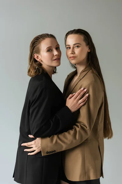 Sensual pareja de lesbianas en negro y beige blazers abrazando y mirando a la cámara aislada en gris - foto de stock