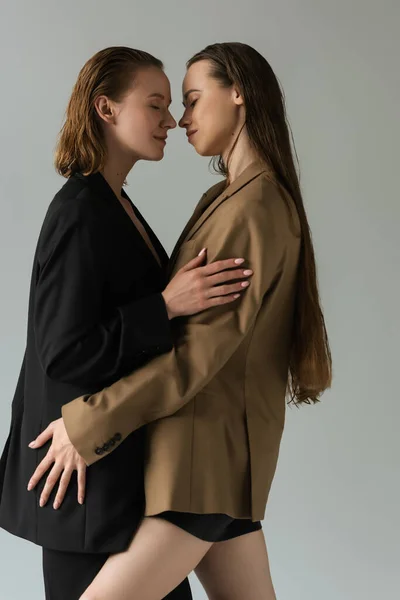 Vue latérale de femmes lesbiennes en blazers beige et noir debout face à face avec les yeux fermés isolés sur gris — Photo de stock