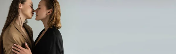 Вид збоку красивої лесбійської пари в куртках, що стоять обличчям до обличчя ізольовані на сірому, банер — стокове фото