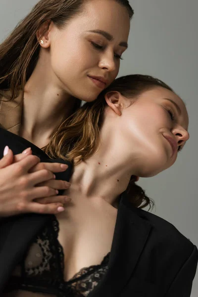 Low angle view of young lesbian woman near seductive girlfriend in black bra and jacket isolated on grey — Stock Photo