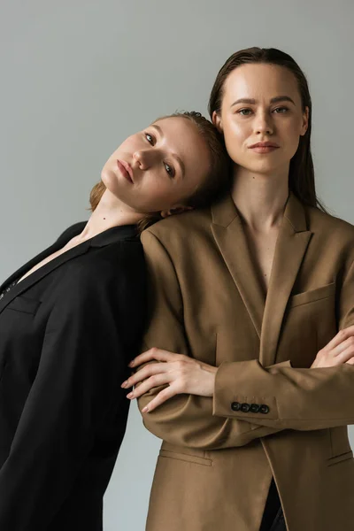 Young woman in black blazer leaning on lesbian girlfriend standing with crossed arms isolated on grey — Stock Photo