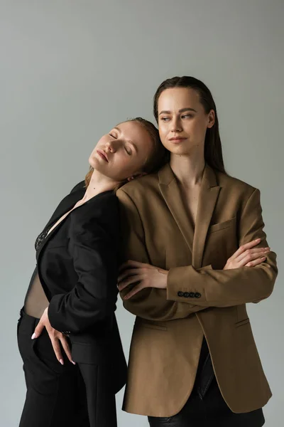 Sensual lesbian woman with closed eyes leaning on girlfriend standing with crossed arms isolated on grey — Stock Photo