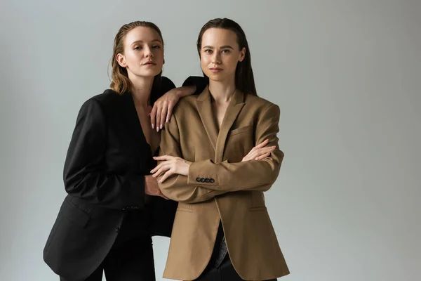 Lesbian woman in black blazer leaning on girlfriend standing with crossed arms and looking at camera isolated on grey — Stock Photo