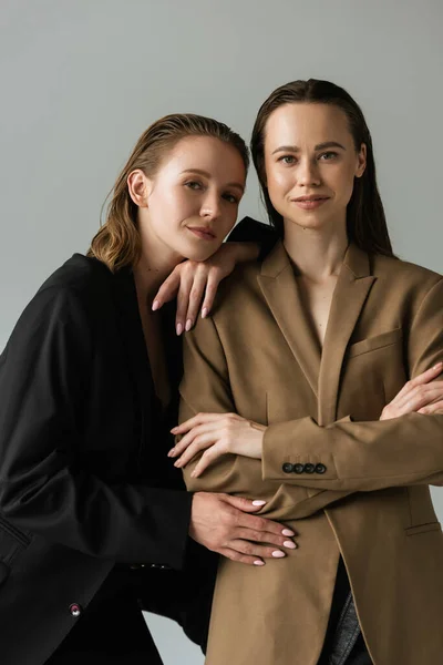 Jeune femme penché sur lesbienne copine debout avec les bras croisés et regardant caméra isolé sur gris — Photo de stock