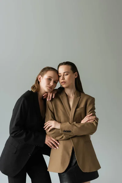 Lesbische Frau im beigen Blazer steht mit verschränkten Armen neben hübscher Freundin isoliert auf grau — Stock Photo