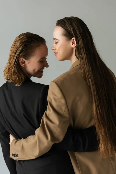 Back view de casal lésbico em blazers pretos e bege sorrindo e abraçando isolado em cinza — Fotografia de Stock