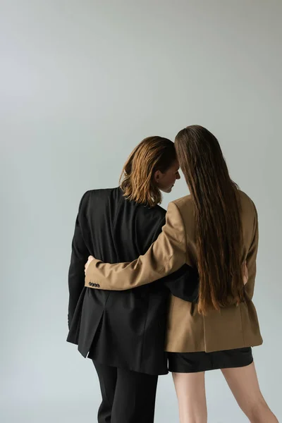 Back view of woman with long hair hugging lesbian girlfriend in black blazer isolated on grey — Stock Photo