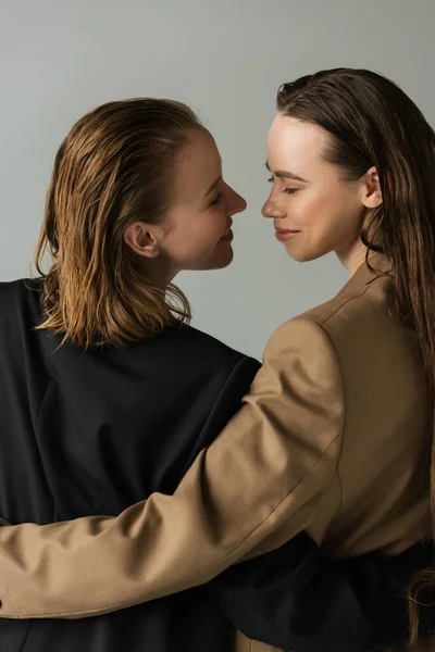 Cheerful lesbian couple hugging and smiling at each other isolated on grey — Stock Photo