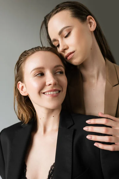 Mulher em blazer preto sorrindo perto da namorada morena concurso isolado em cinza — Fotografia de Stock