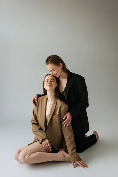 Happy lesbian woman in black suit embracing girlfriend sitting with closed eyes on grey background — Stock Photo