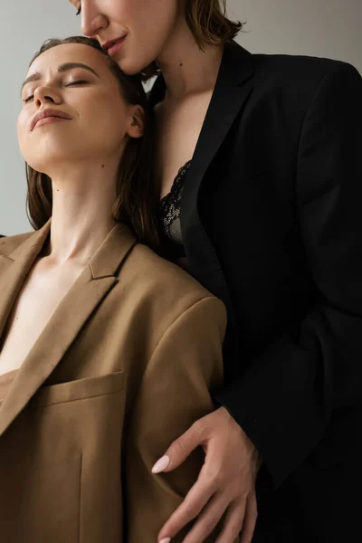 Woman in beige blazer standing with closed eyes near lesbian girlfriend embracing her isolated on grey — Stock Photo