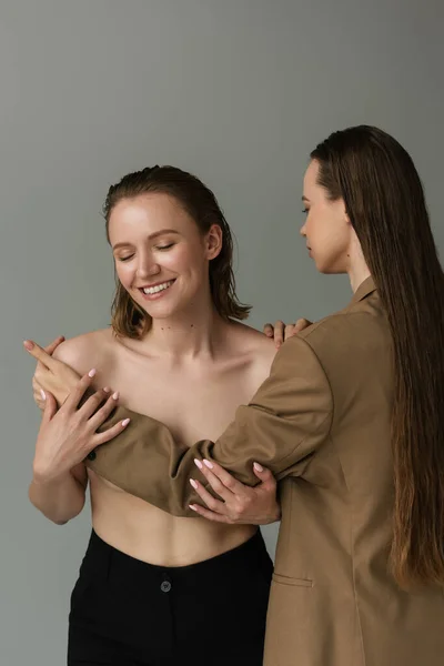 Jeune lesbienne femme souriant près petite amie couvrant son sein isolé sur gris — Photo de stock