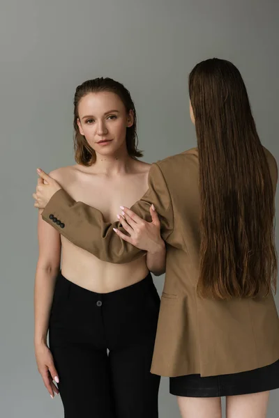 Young woman looking at camera near long haired girlfriend covering her breast isolated on grey — Stock Photo