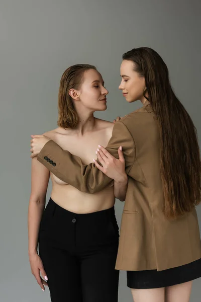 Улыбающаяся лесбиянка с длинными волосами, покрывающая грудь молодой и сексуальной девушки, изолированной на серой — стоковое фото