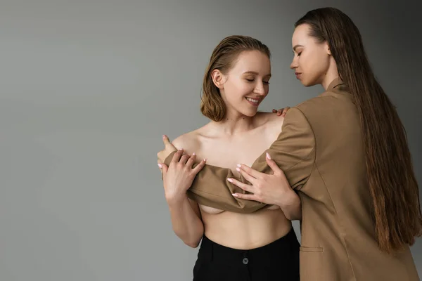 Femme aux cheveux longs en blazer beige couvrant le sein d'une petite amie lesbienne heureuse isolée sur gris — Photo de stock