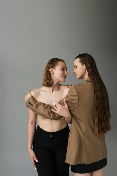 Длинноволосая женщина в бежевом блейзере, покрывающая грудь молодой лесбиянки, изолированной на серой — стоковое фото