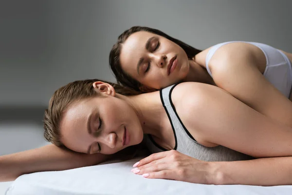 Smiling woman with closed eyes relaxing on bed near brunette girlfriend on grey background — Stock Photo