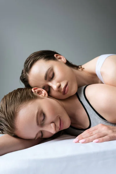 Bella donna lesbica sorridente con gli occhi chiusi mentre sdraiato sul letto vicino bruna fidanzata isolato su grigio — Foto stock