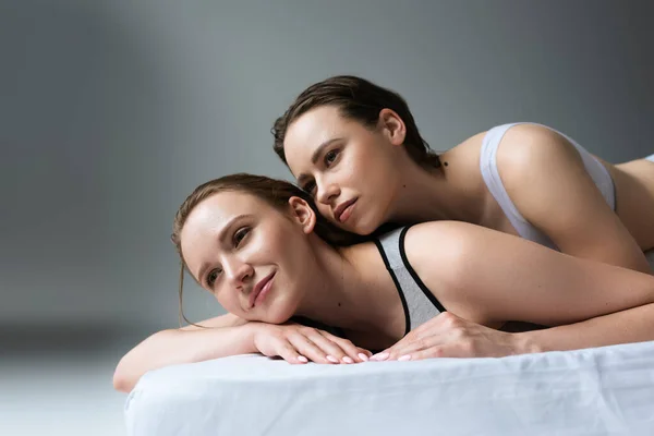 Lächelnde lesbische Frau schaut weg, während sie es sich auf dem Bett neben brünetter Freundin auf grauem Hintergrund gemütlich macht — Stockfoto