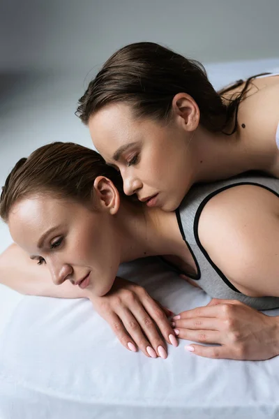 Sexy brunette femme près de jeune lesbienne copine couché sur blanc literie sur fond gris — Photo de stock