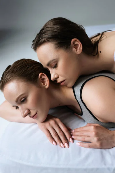 Sexy brunette woman near young lesbian girlfriend lying on white bedding on grey background — Stock Photo