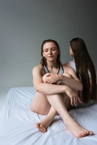 Young lesbian woman sitting on bed with crossed legs near long haired girlfriend isolated on grey — Stock Photo