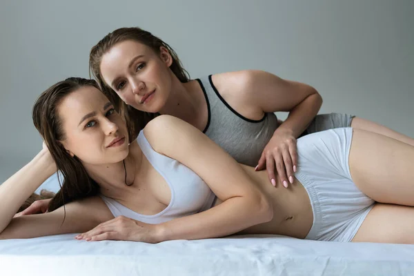 Sexy couple of lesbians lying on bed in underwear and looking at camera isolated on grey — Stock Photo