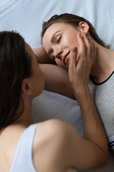 Vista de alto ângulo de mulher lésbica borrada tocando rosto de namorada deitada em roupa de cama branca com olhos fechados — Fotografia de Stock