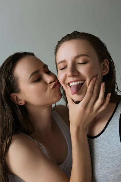 Young woman pouting lips and touching face of lesbian girlfriend sticking out tongue isolated on grey — Stock Photo