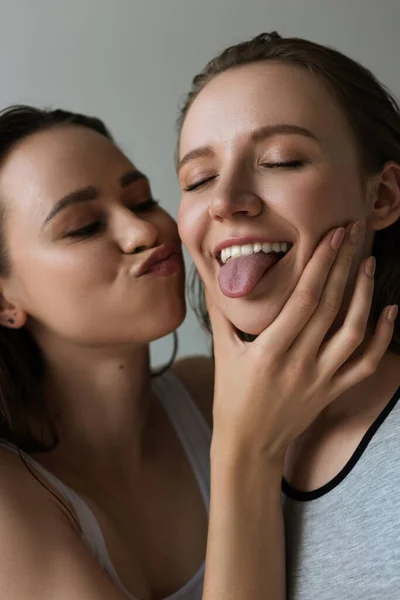 Fröhliche Frau mit geschlossenen Augen, die Zunge herausstreckt, neben lesbischer Freundin schmollende Lippen isoliert auf grau — Stockfoto