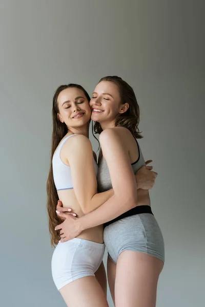 Feliz mismo sexo lesbianas pareja en ropa interior abrazando con cerrado ojos aislado en gris — Stock Photo