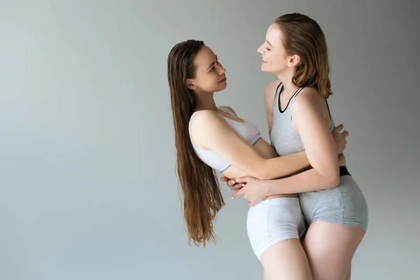 Vista lateral de lesbianas jóvenes y felices en ropa interior abrazándose y mirándose aisladas en gris - foto de stock