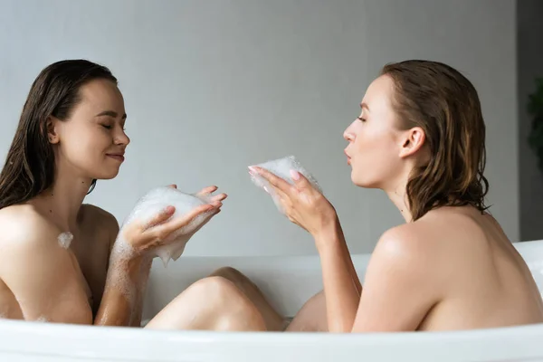 Side view of naked lesbian couple having fun with soap foam while taking bath together — Stock Photo