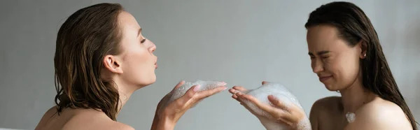 Vista lateral de mujer joven con los ojos cerrados divirtiéndose con novia lesbiana y espuma de jabón en el baño, pancarta - foto de stock