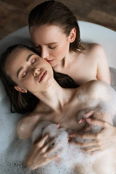 Вид сверху на молодую женщину, обнимающую грудь подруги-лесбиянки во время принятия ванны с закрытыми глазами — стоковое фото