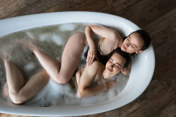 Вид сверху на сексуальных лесбиянок с закрытыми глазами, принимающих ванну с мыльными пузырями дома — стоковое фото
