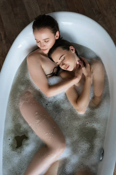 Vista superior de pareja lesbiana desnuda con los ojos cerrados tomando baño de burbujas en casa - foto de stock