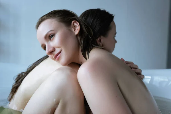 Feliz lesbiana abrazando con novia en bañera y mirando hacia otro lado - foto de stock